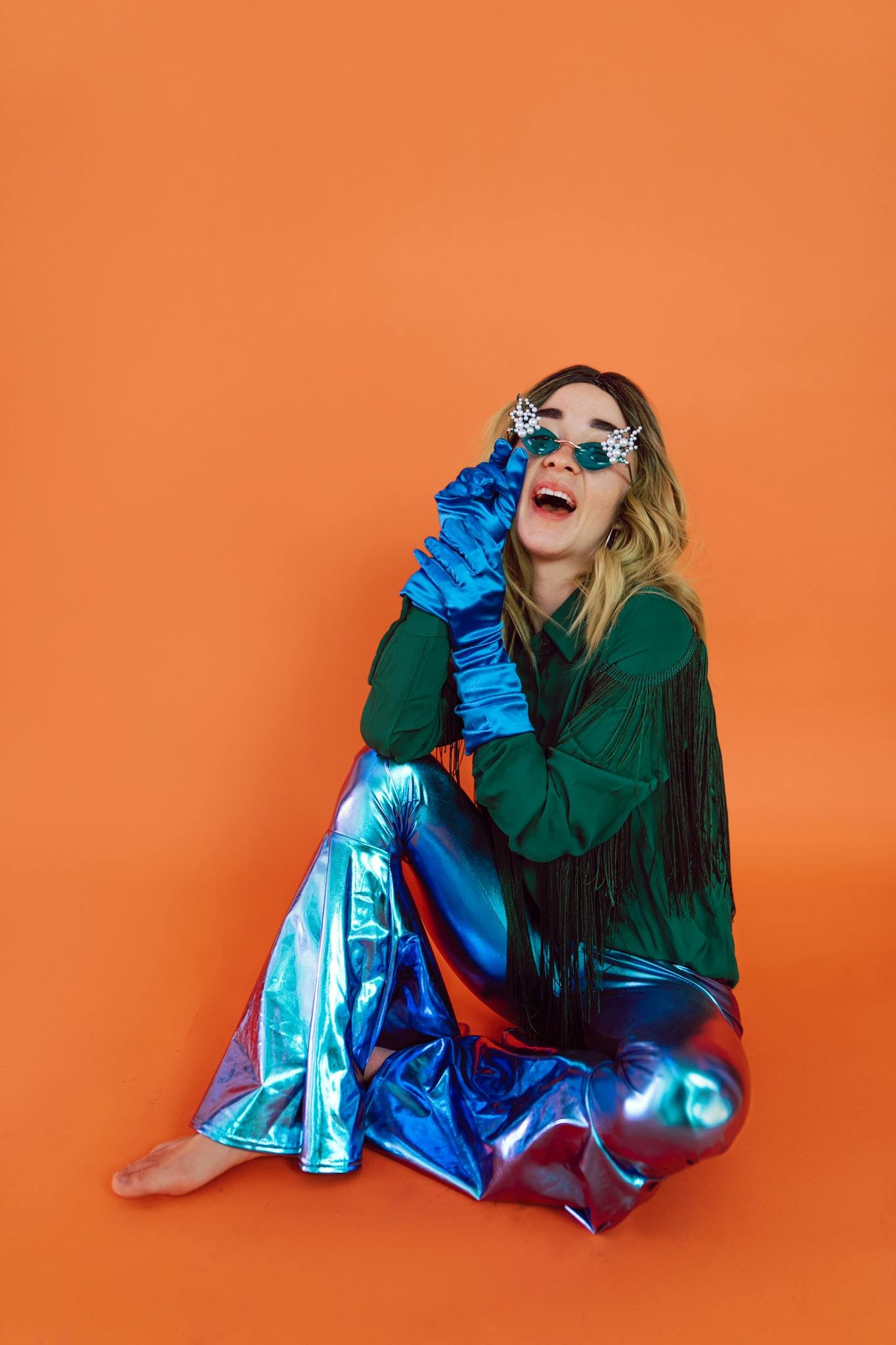 Colorful portrait of a woman in metallic pants and playful accessories against an orange backdrop.