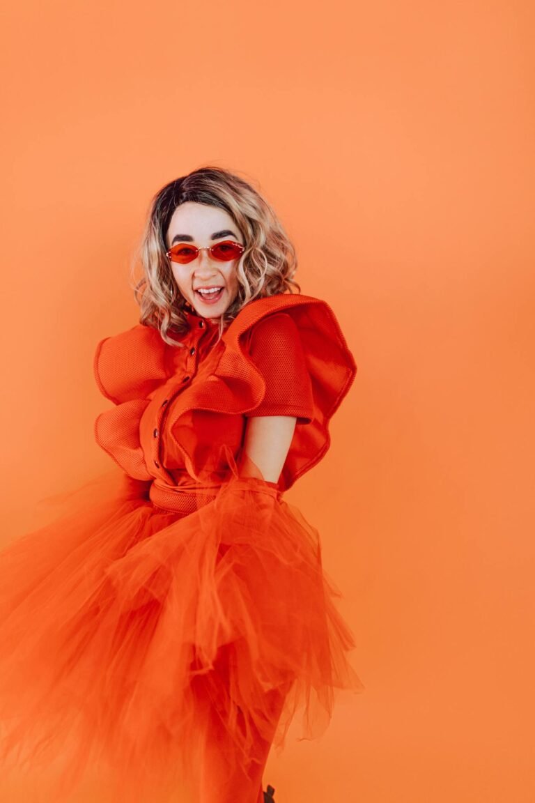 Dynamic portrait of a stylish woman in a vibrant orange dress and red glasses against an orange backdrop.