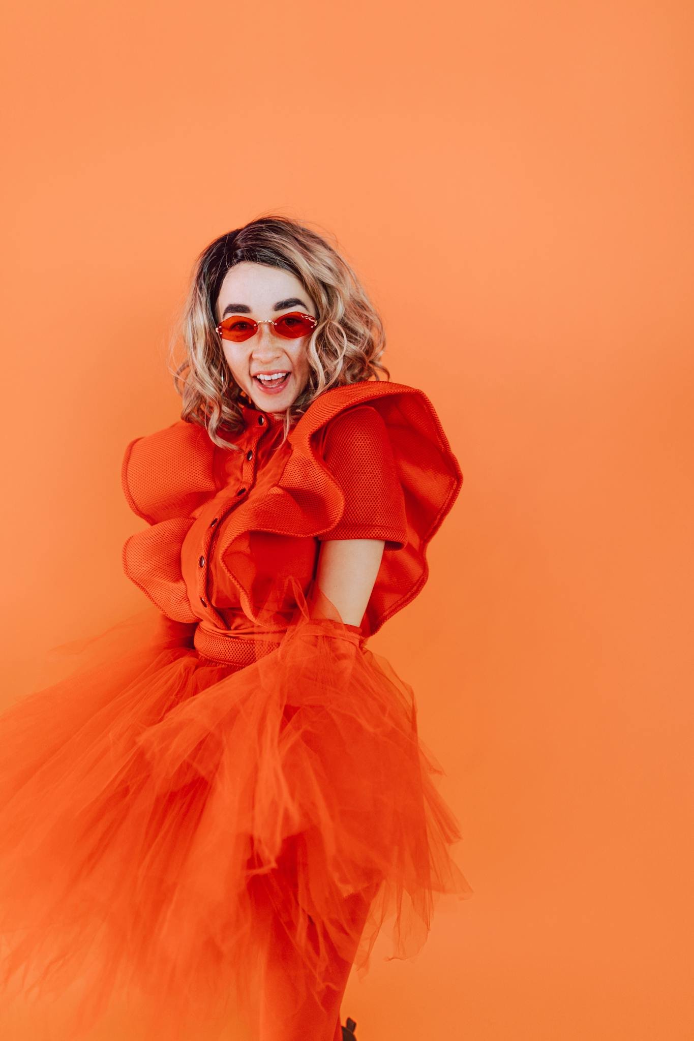 Dynamic portrait of a stylish woman in a vibrant orange dress and red glasses against an orange backdrop.