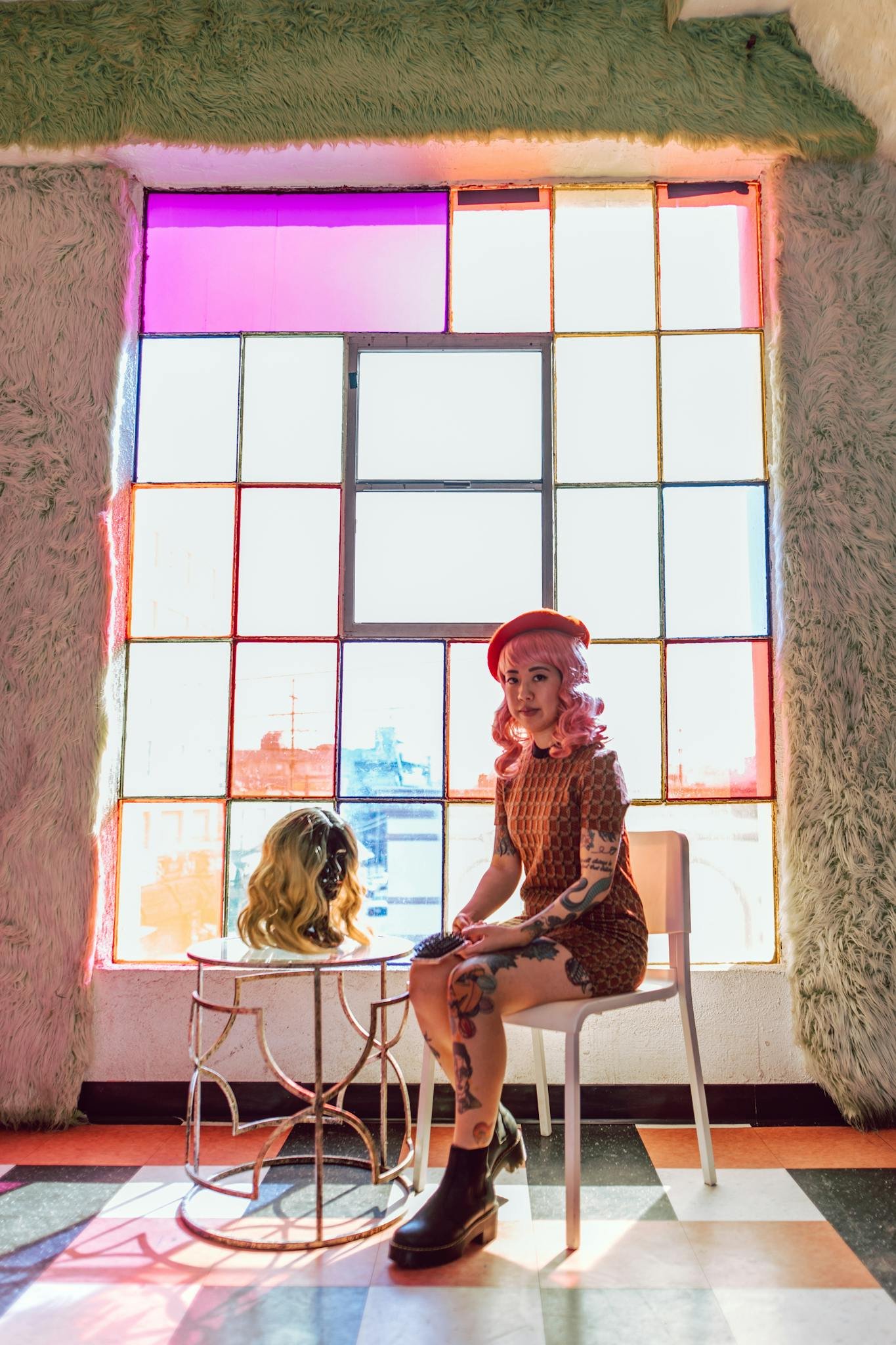Stylish woman with colorful hair and tattoos seated indoors next to a wig on display. Vibrant atmosphere.
